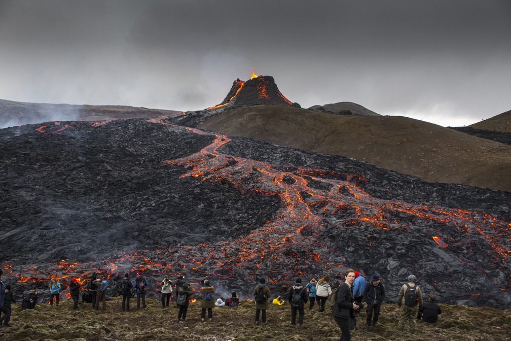 Curiosos assistem e tiram fotos de vulcão na Islândia enquanto a lava flui da erupção na península de Reykjanes na terça-feira (23) — Foto: Marco Di Marco/AP